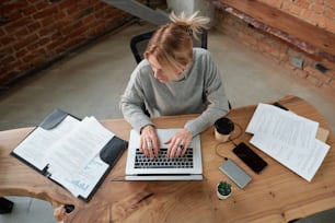 Above view of female manager in sweater typing on laptop while analyzing papers in office