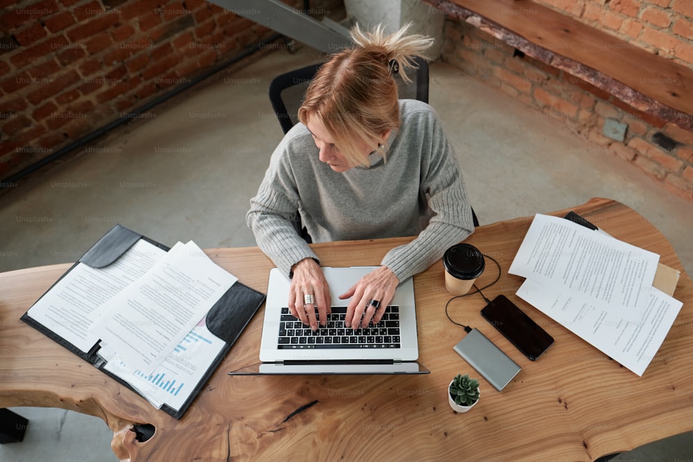 Vista sopra della manager femminile in maglione che digita sul computer portatile mentre analizza i documenti in ufficio