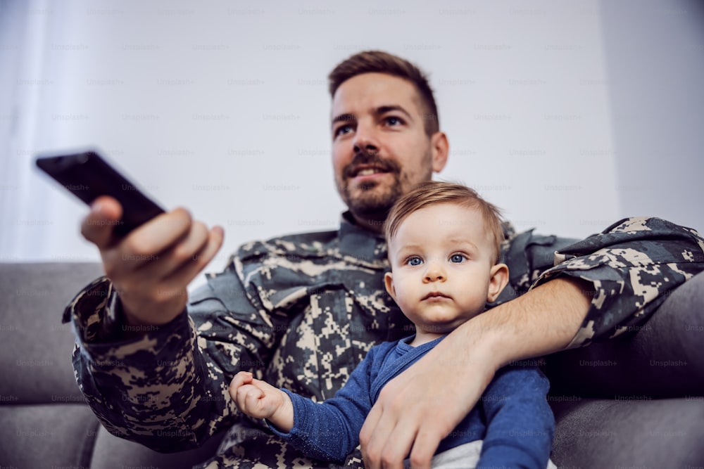 Young bearded brave soldier spending quality time with his beloved son. They are watching television. Man having remote control in hands.