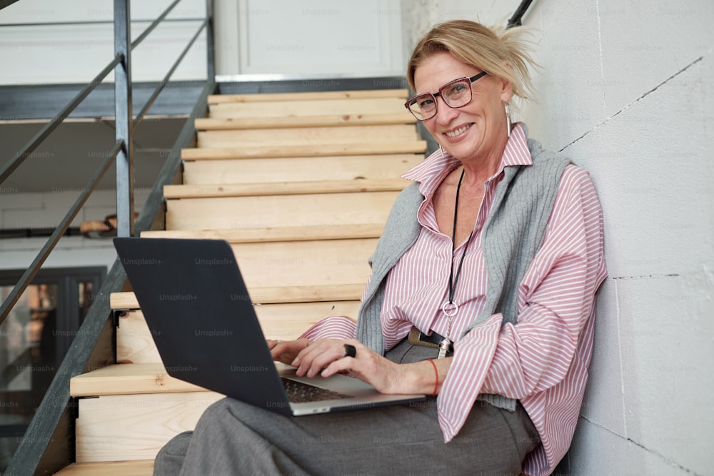 Ritratto di donna d'affari matura allegra seduta sulle scale e utilizzando il computer portatile durante la preparazione del rapporto di vendita