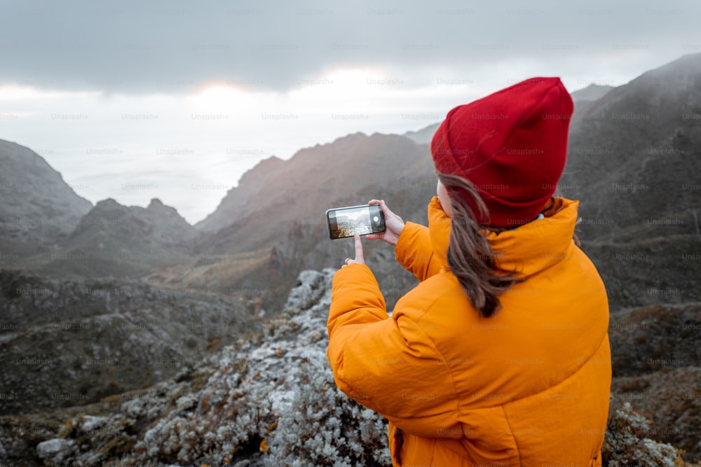 Young woman dressed in bright jacket and hat photographing on phone breathtaking views on the mountain range under the clouds, traveling on Tenerife island, Spain
