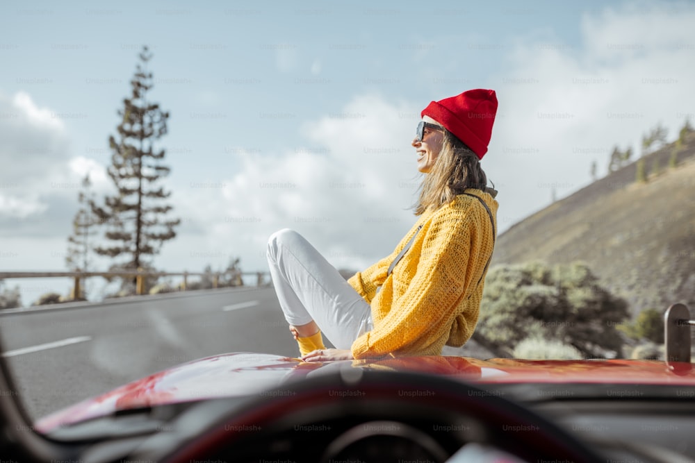Ritratto di stile di vita di una donna spensierata vestita casualmente con maglione luminoso e cappello seduta sul cofano dell'auto, godendo di un viaggio sulla strada di montagna, vista attraverso il parabrezza