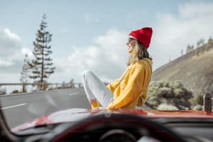 Retrato de estilo de vida de una mujer despreocupada vestida casualmente con suéter brillante y sombrero sentada en el capó del automóvil, disfrutando del viaje por carretera en la carretera de montaña, vista a través del parabrisas