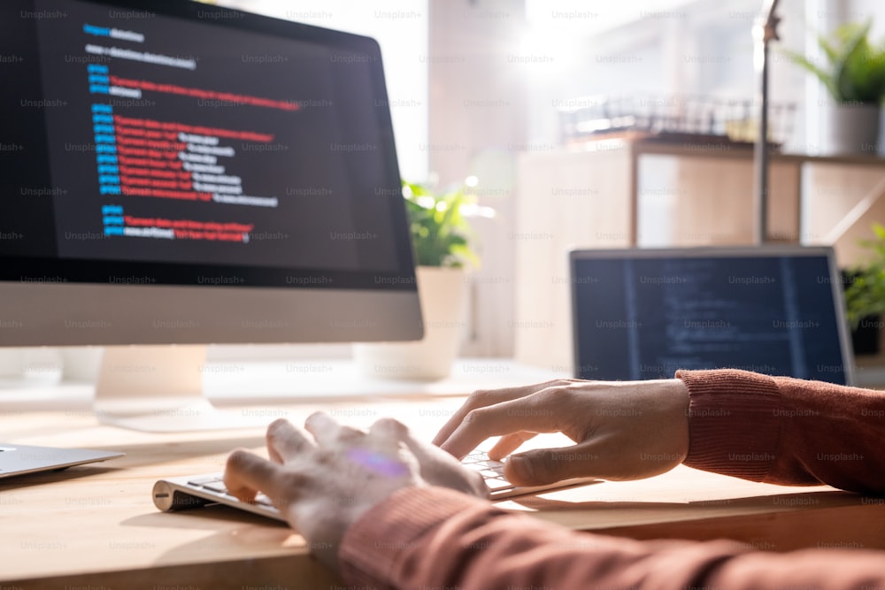 Hands of young contemporary programmer over keypad during work over new software in front of computer screen in office