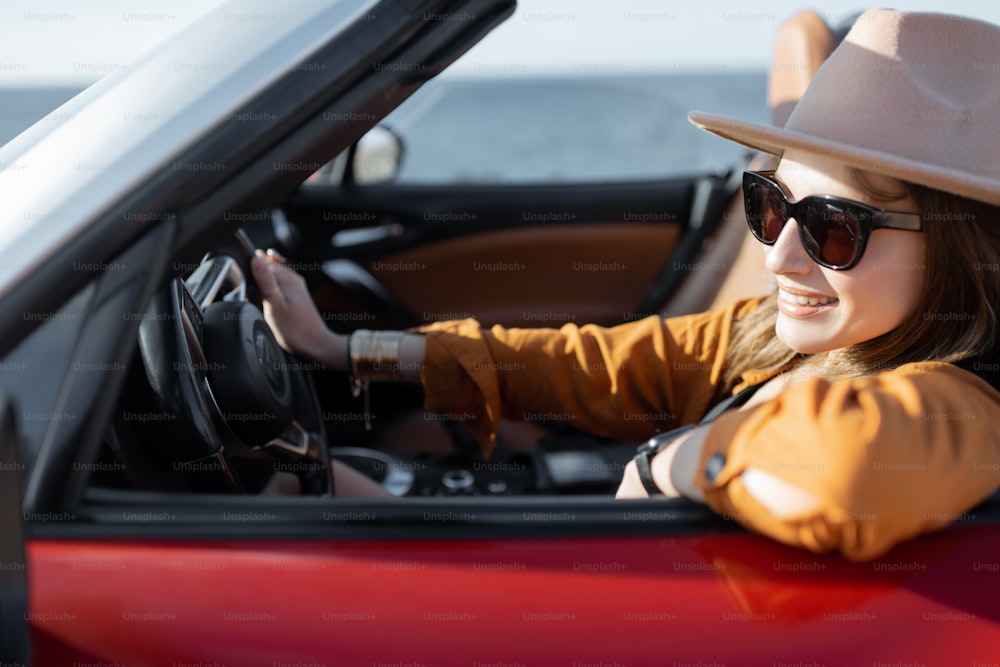 Mulher elegante jovem no chapéu dirigindo carro conversível perto do oceano, desfrutando de férias de verão enquanto viaja na ilha. Conceito de viagem despreocupada e horário de verão