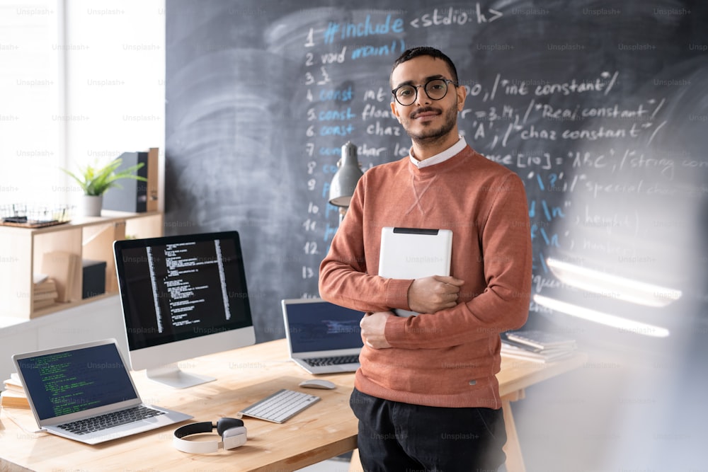 Young casual software developer with digital tablet standing by table in front of camera with blackboard on background