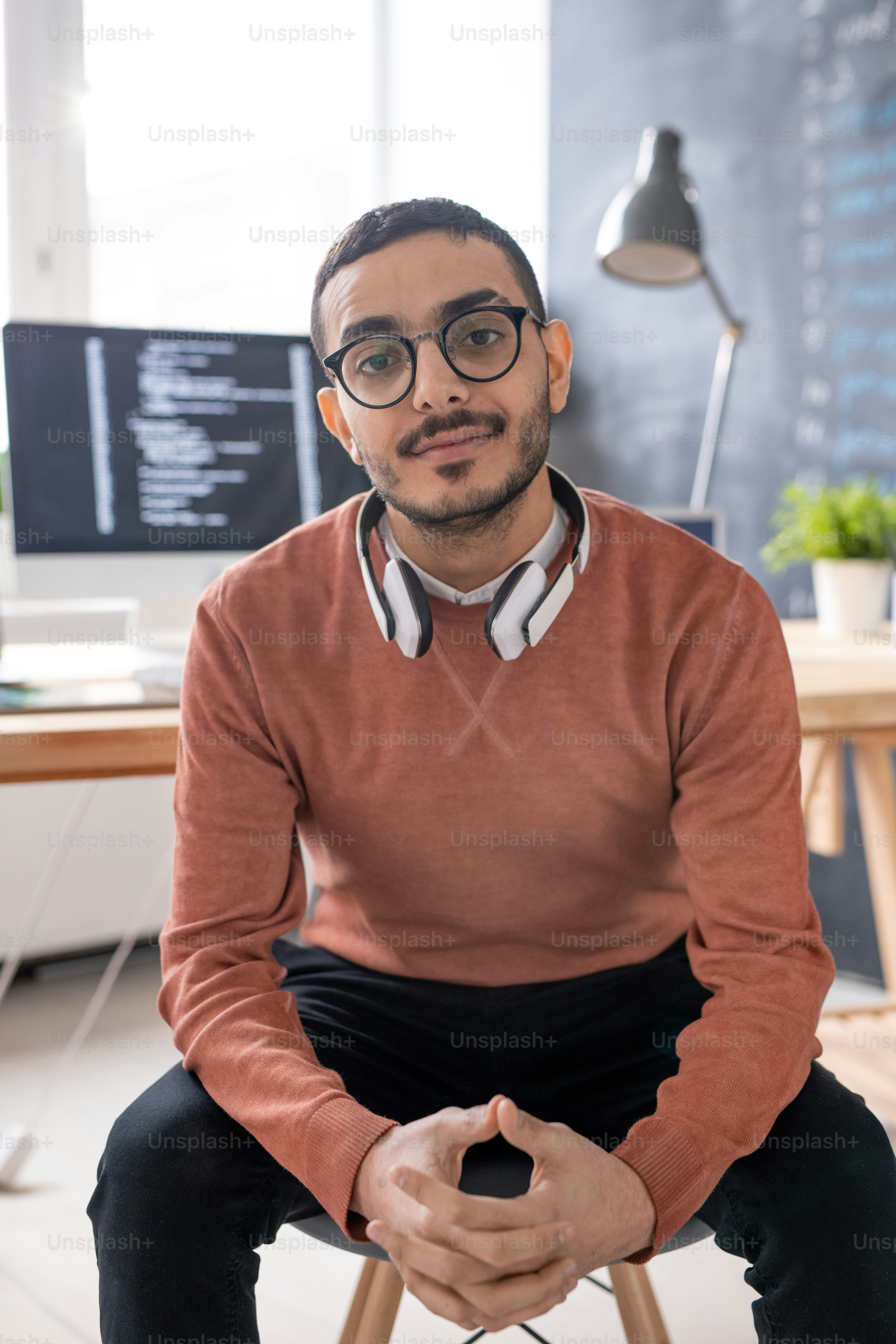 Programmer kasual muda dengan headphone menatap Anda sambil duduk di depan kamera dengan latar belakang tempat kerja