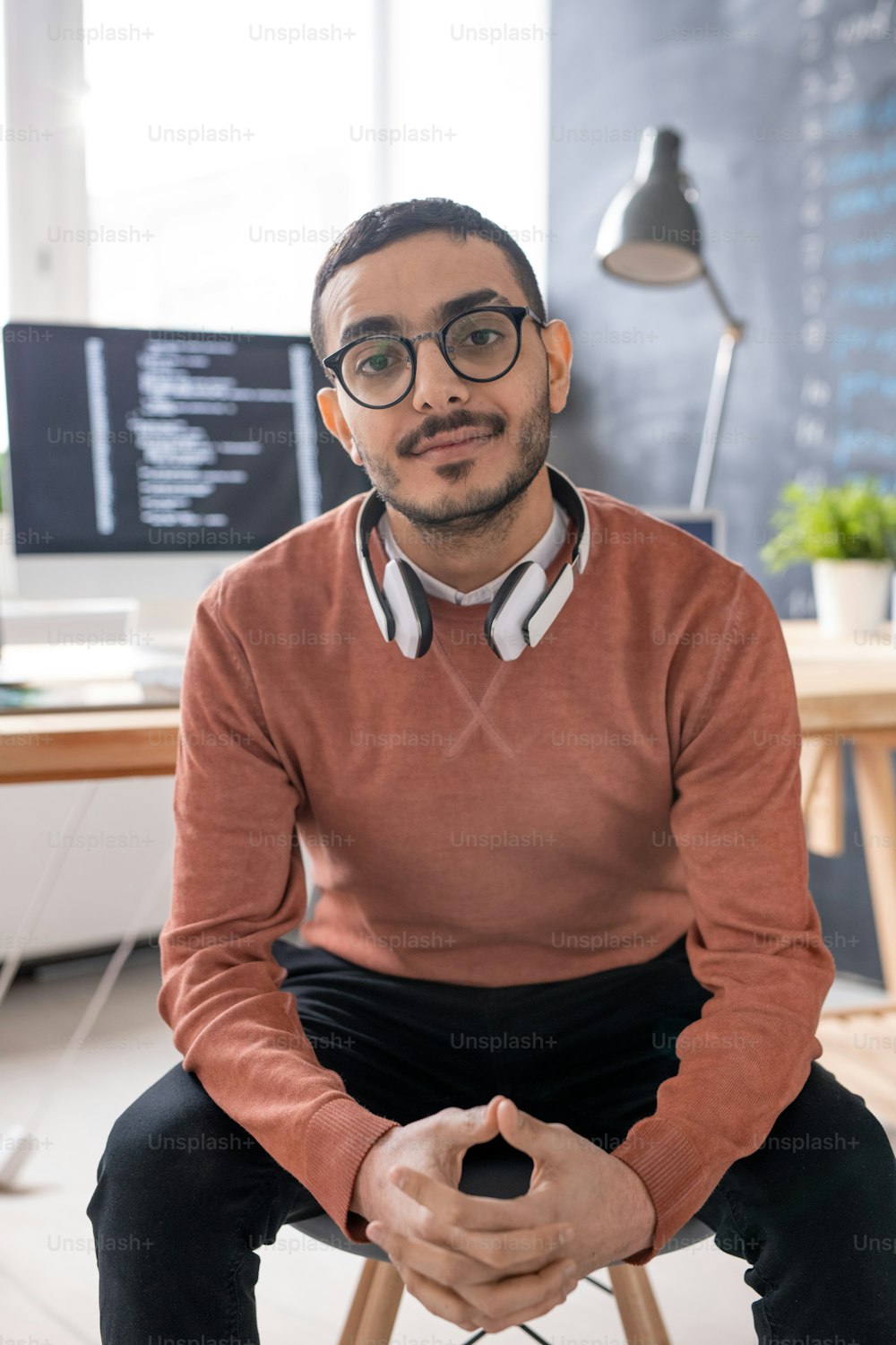 Young casual programmer with headphones looking at you while sitting in front of camera with workplace on background