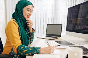 Happy young Asian freelance software developer talking on phone with client and taking notes in planner