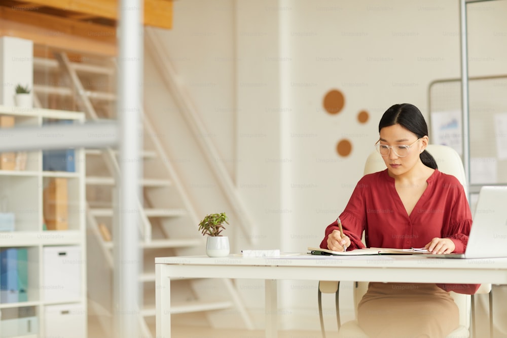 Retrato de una exitosa empresaria asiática escribiendo en el escritorio en el interior de la oficina blanca moderna, espacio de copia
