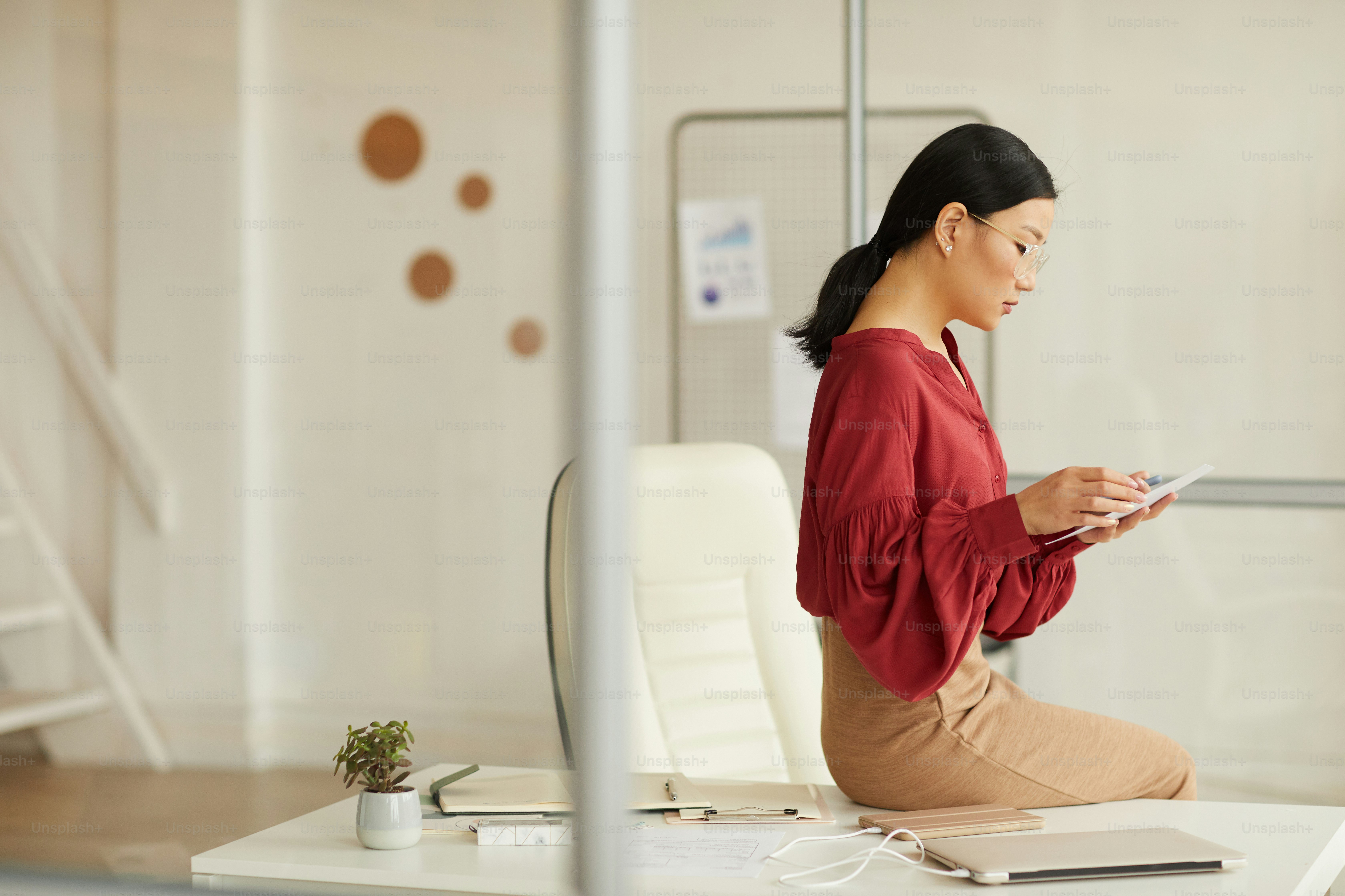 Side view portrait of elegant Asian businesswoman sitting on desk and using digital tablet in modern white office, copy space