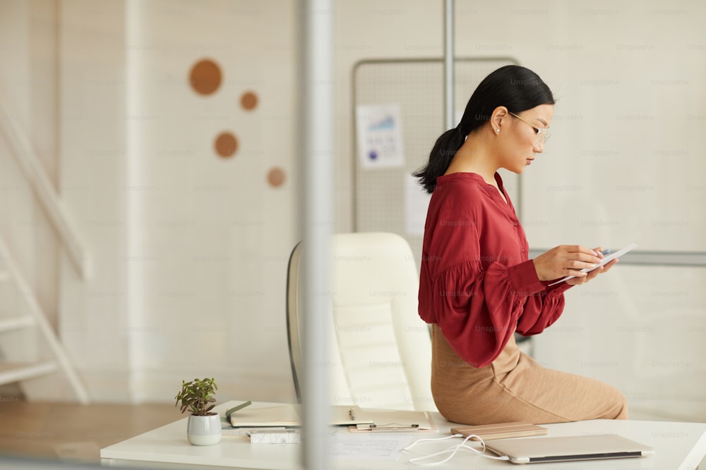 Retrato de vista lateral de una elegante mujer de negocios asiática sentada en el escritorio y usando una tableta digital en una oficina blanca moderna, espacio de copia
