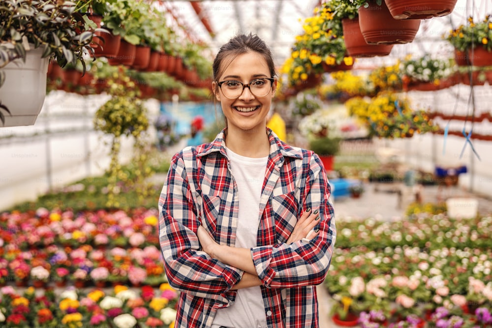 Young smiling successful female entrepreneur standing in nursery garden with arms folded and looking at camera. In background are all sorts of colorful flowers.