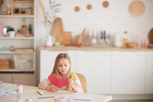 Weitwinkelporträt eines niedlichen blonden Mädchens, das eine herzförmige Karte hält, während es am Tisch in gemütlichem Wohnraum sitzt