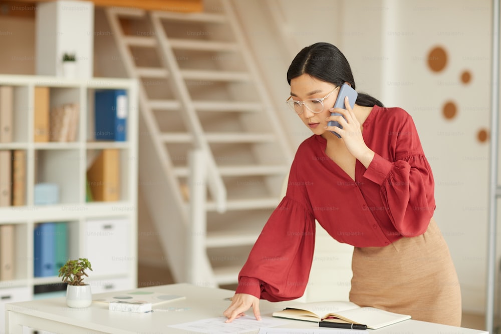 Retrato de una elegante empresaria asiática que revisa el planificador mientras habla por teléfono en una moderna oficina blanca, espacio de copia