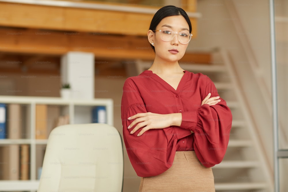 Ritratto in vita di donna asiatica di successo in piedi con le braccia incrociate mentre posa in ufficio moderno, spazio copia