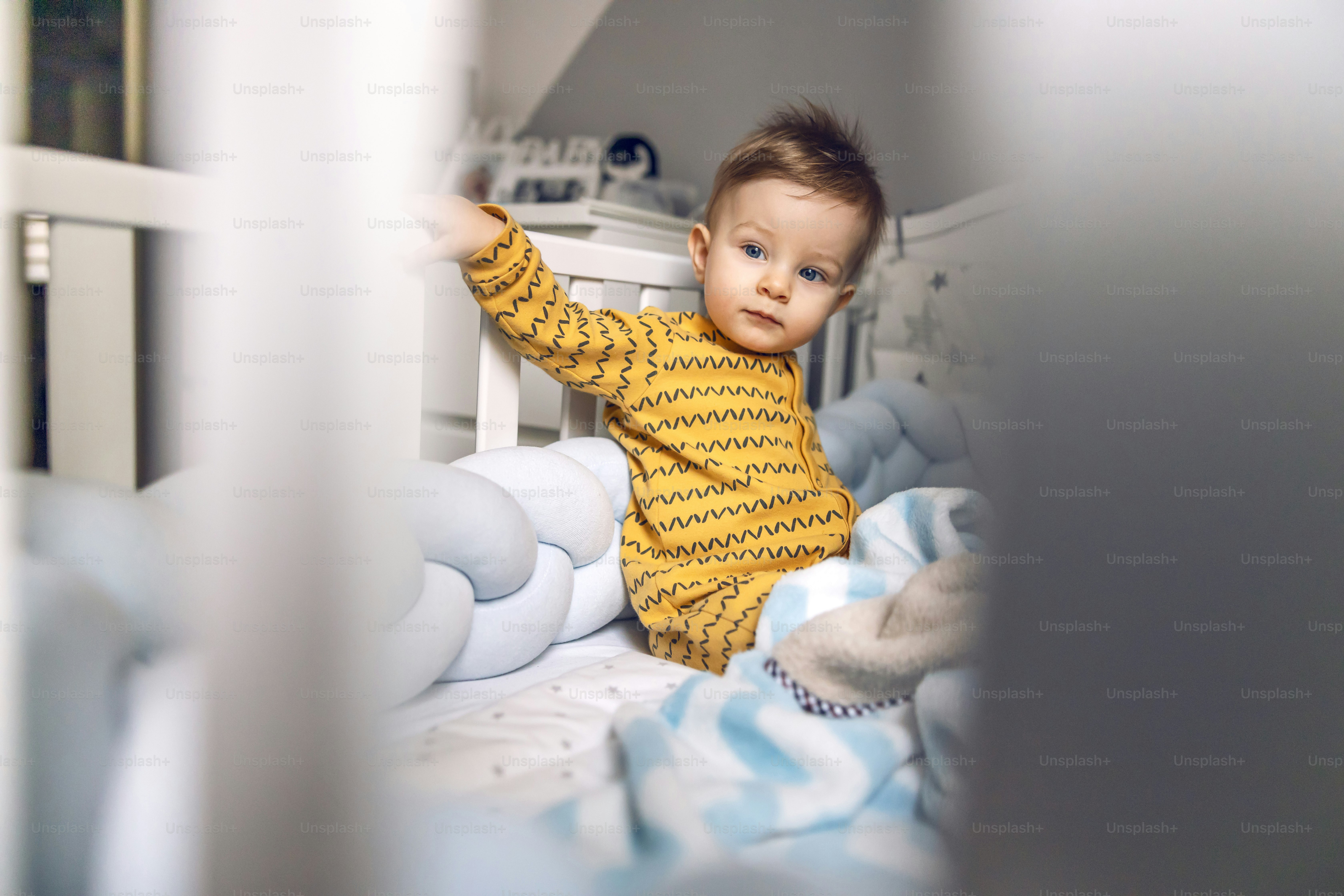 Cute little blond baby boy sitting in his crib in the morning with serious facial expression.