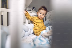 귀여운 금발의 아기가 진지한 표정으로 아침에 아기 침대에 앉아 있다.