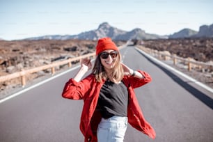 晴れた日に火山の谷の真ん中にある美しい道を歩く赤い服を着たスタイリッシュな若い女性のライフスタイルポートレート。のんきなライフスタイルと旅行のコンセプト