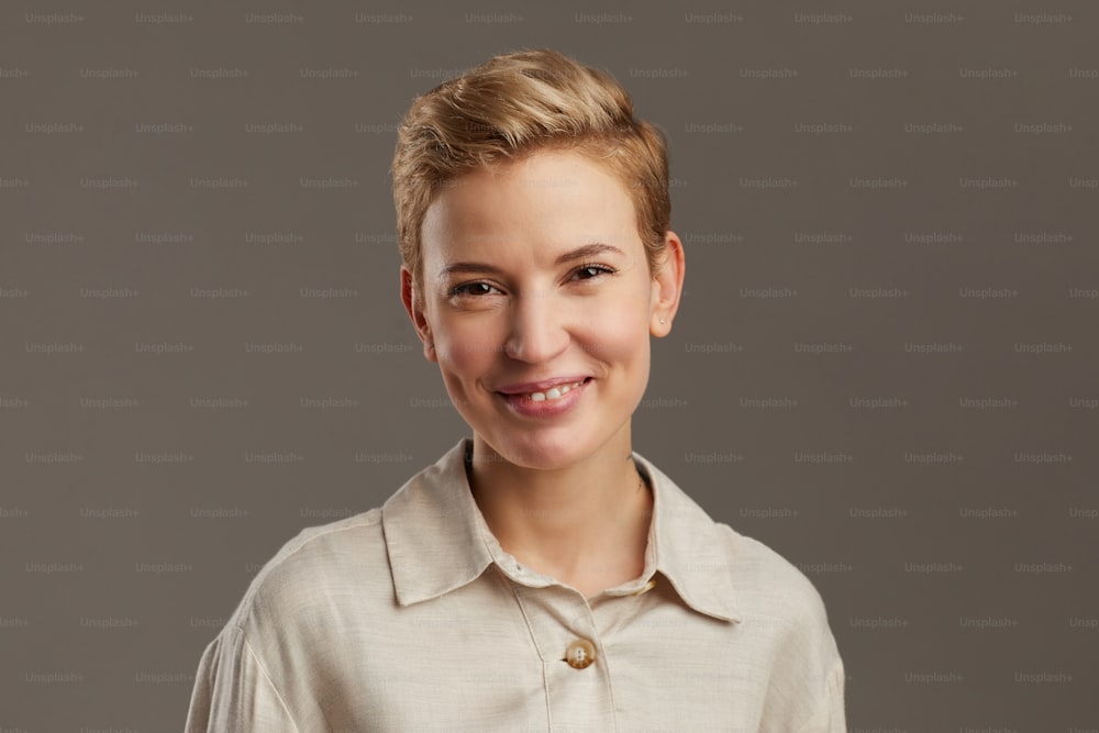 Porträt einer lächelnden jungen Frau mit kurzem Pixie-Haarschnitt, die fröhlich in die Kamera schaut, während sie vor grauem Hintergrund im Studio steht, Kopierraum