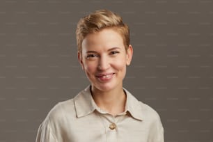Portrait of smiling young woman with short pixie haircut looking cheerfully at camera while standing against grey background in studio, copy space