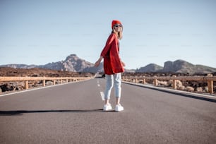 Lifestyle portrait of a young woman stylishly dressed in red walking on the beautiful road in the midst of volcanic valley on a sunny day. Carefree lifestyle and travel concept