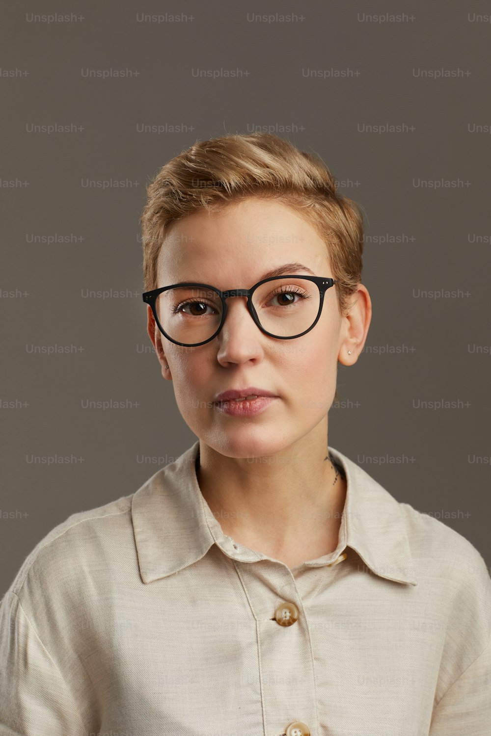 Portrait of modern young woman with short pixie haircut looking at camera while standing against grey background in studio