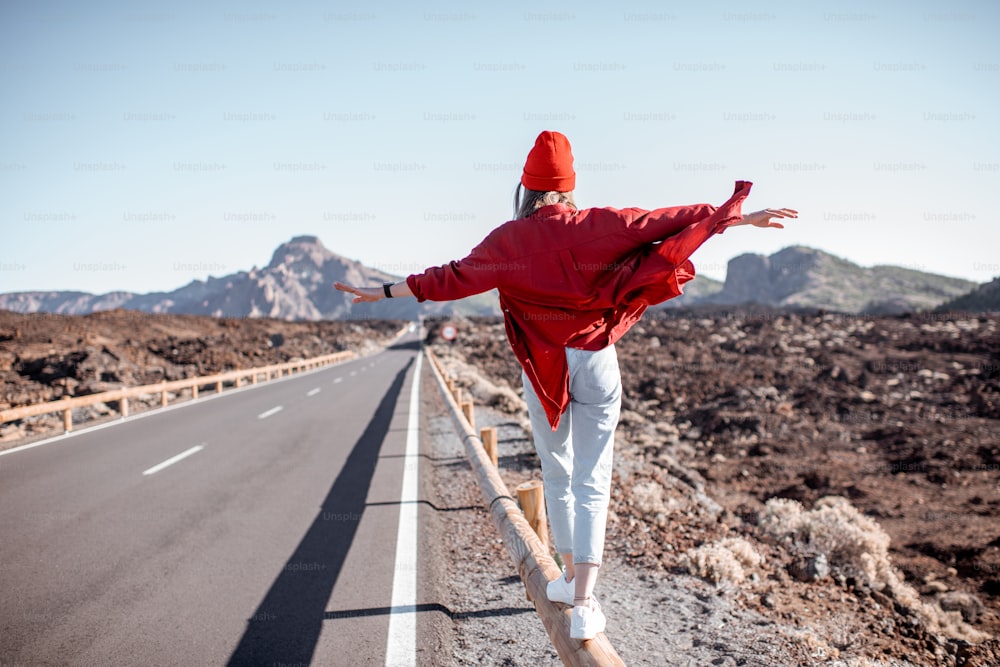 Lifestyle-Porträt einer jungen, stilvoll in Rot gekleideten Frau, die an einem sonnigen Tag inmitten eines Vulkantals auf dem Straßenzaun spazieren geht. Unbeschwertes Lifestyle- und Reisekonzept