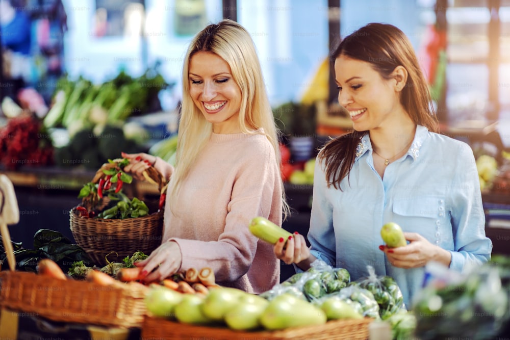 Two attractive smiling best friends with healthy habits choosing vegetables at farmers market. Blonde one holding basket.