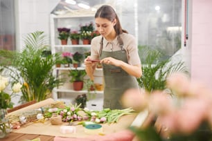 Retrato de cintura para arriba de una joven creativa que toma una foto de teléfono inteligente de flores de tulipán mientras trabaja en una floristería, copia el espacio