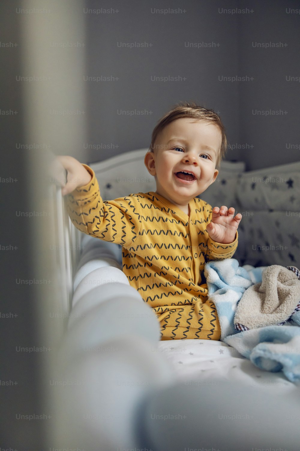 Cheerful smiling adorable blond baby boy sitting in his crib in the morning and waving. 'Good morning mom and dad!'