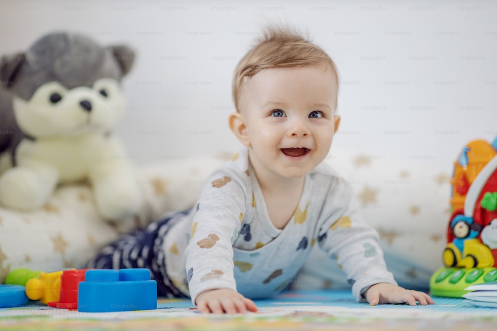 Adorable smiling playful blond little boy with beautiful blue eyes lying on stomach on the floor surrounded by toys.