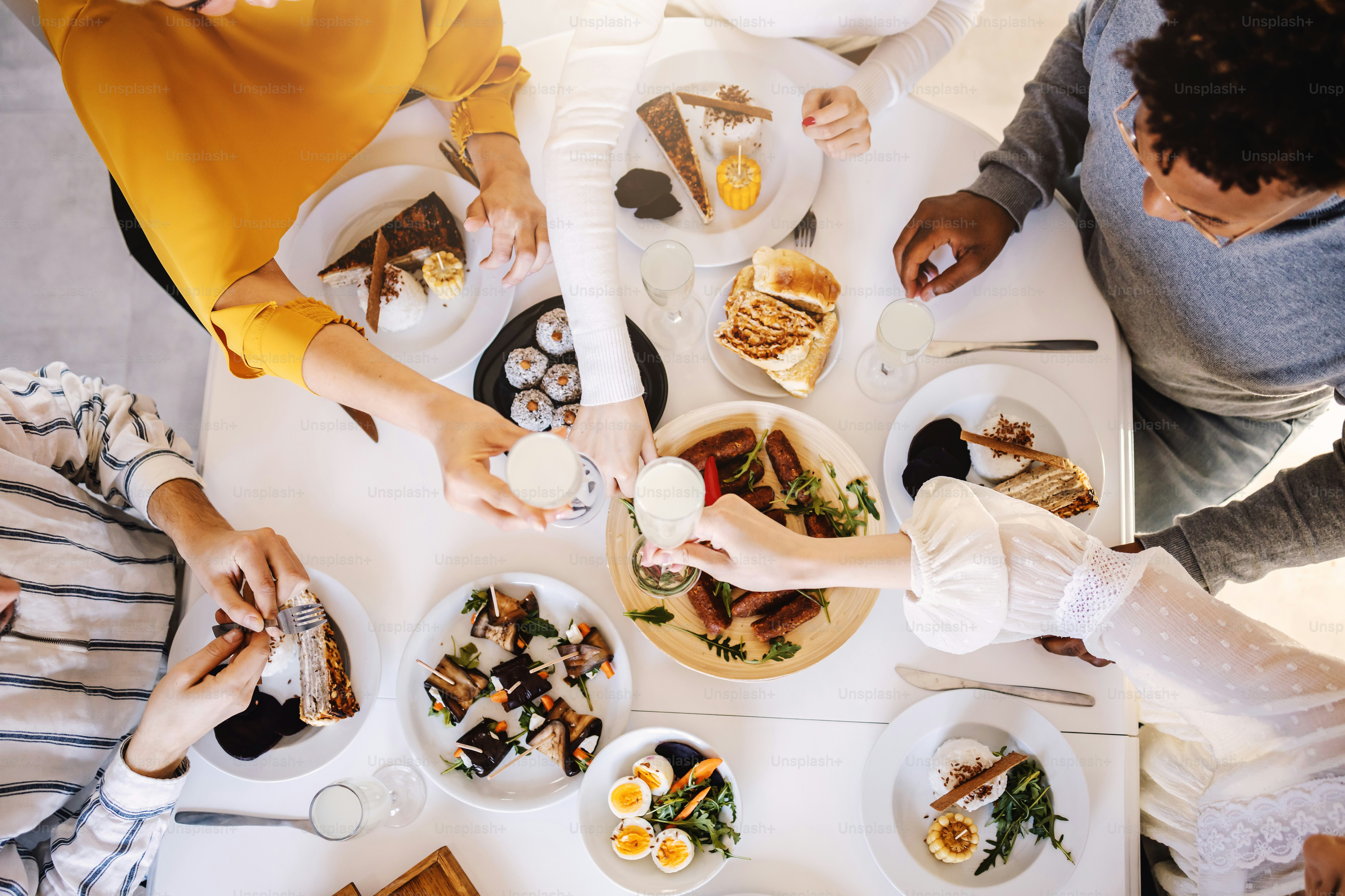 Top view of five multicultural friends sitting at dinning table and having healthy lunch. Women are toasting with fresh lemonade while men eating food.