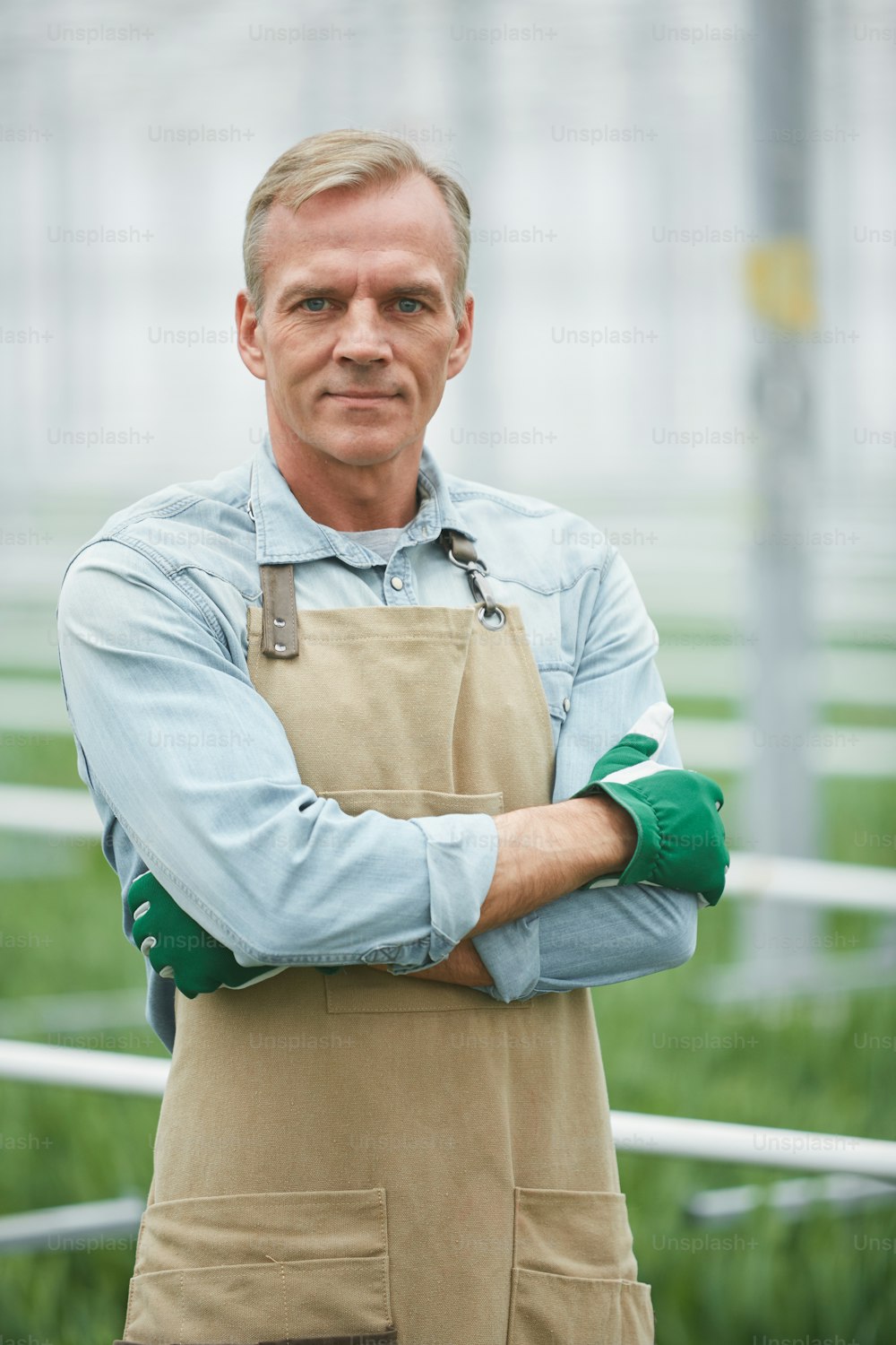 Waist up portrait of mature male worker smiling at camera while standing with arms crossed in industrial greenhouse