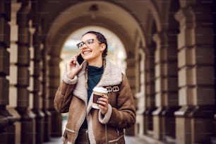 웃고 있는 패셔너블한 10대 소녀가 시내의 오래된 건물 앞에 서서 커피와 함께 일회용 컵을 들고 전화 통화를 하고 있다.