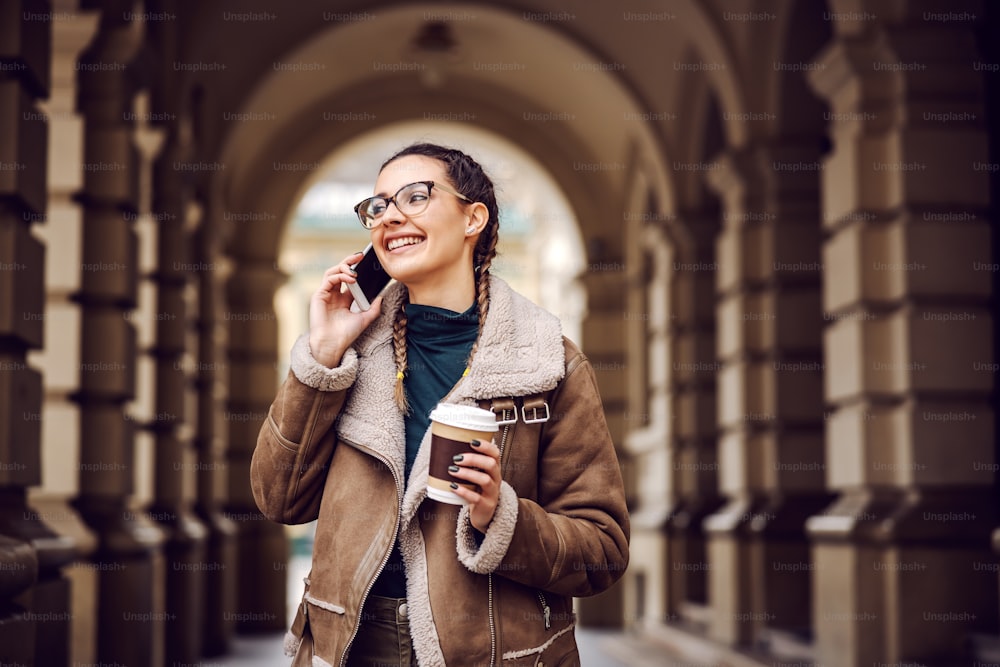 Ragazza adolescente sorridente alla moda in piedi di fronte al vecchio edificio del centro, tenendo in mano una tazza usa e getta con caffè e conversando al telefono.