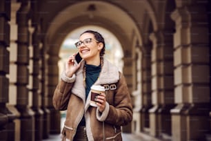 Smiling fashionable teenage girl standing in front of old building downtown, holding disposable cup with coffee and having phone conversation.