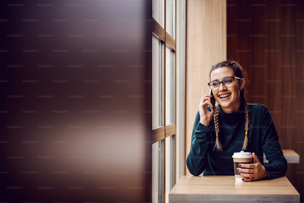 Menina alegre e sorridente sentada no refeitório, conversando por telefone e segurando copo descartável com café fresco.