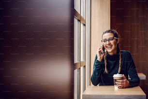 Cheerful smiling young girl sitting in cafeteria, having phone conversation and holding disposable cup with fresh coffee.