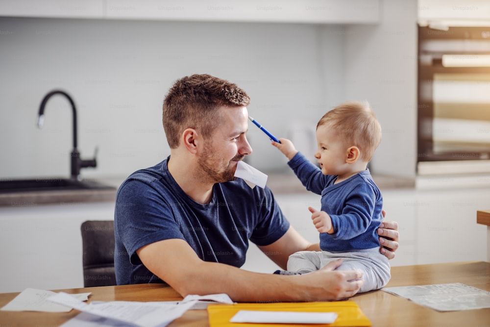 Young attractive bearded father sitting at dinning table and playing with his son. He is holding bill in his mouth. Son sitting on table and trying to help him about finances.