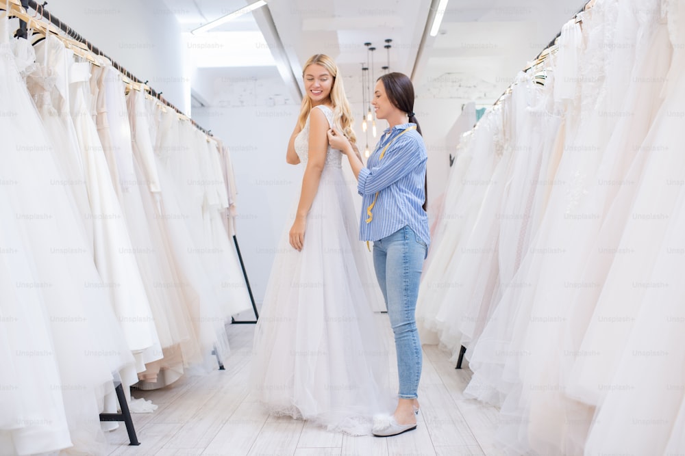 Female shop assistant helping young woman to fasten wedding dress in modern bridal store, horizontal shot