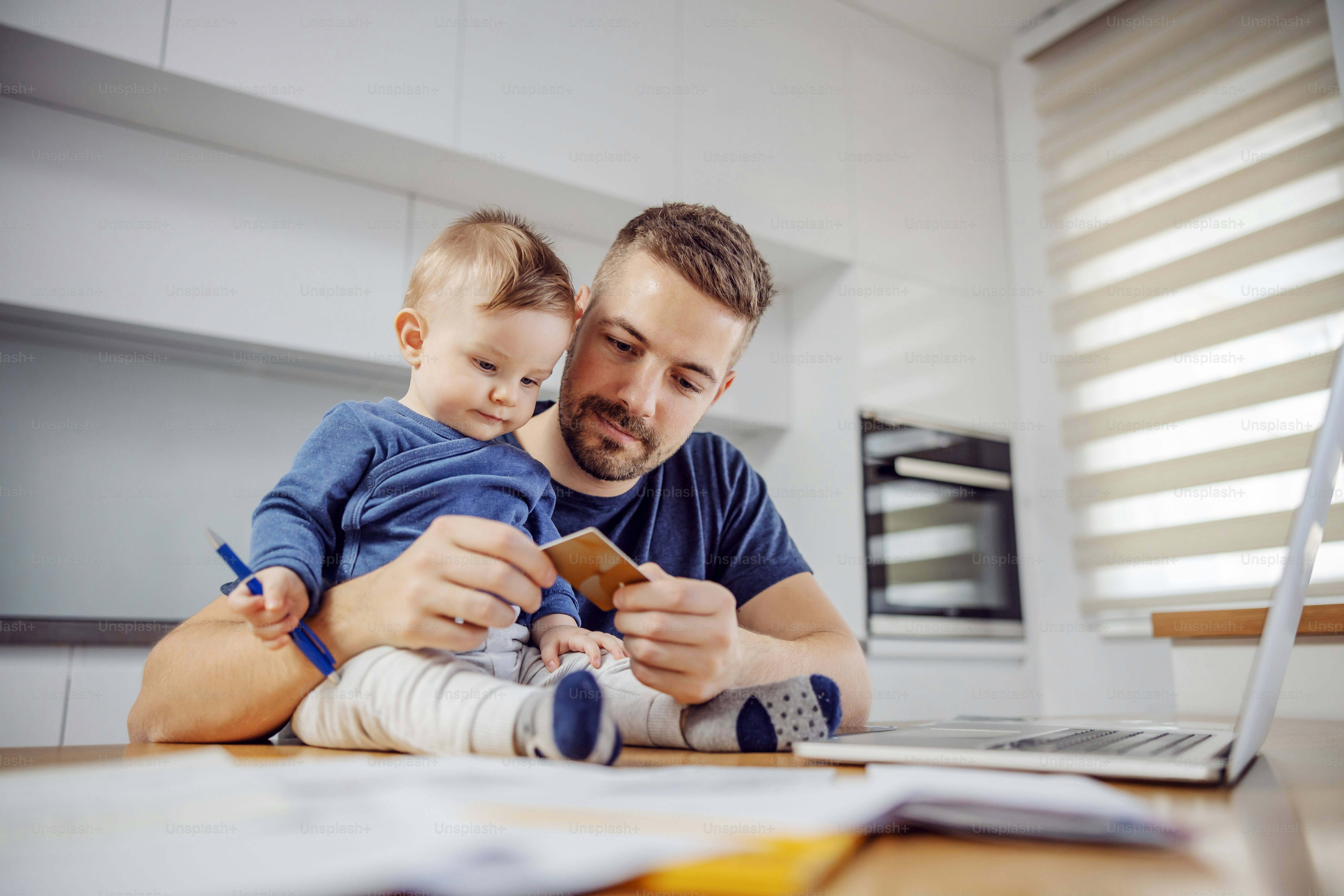 Young attractive bearded father sitting at dinning table with his beloved adorable son and paying bills online. His boy holding pen and trying to help him.