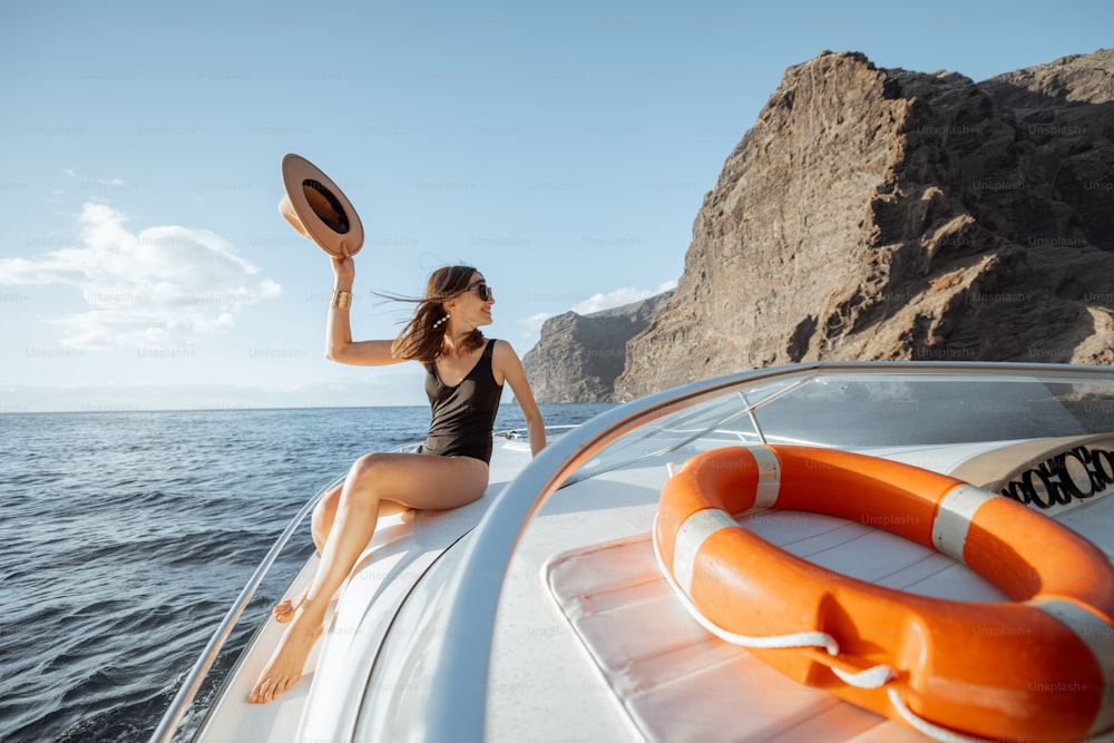 Mulher de maiô e chapéu de sol desfrutando de viagem oceânica, navegando em um iate perto da costa rochosa de tirar o fôlego em um pôr do sol. Conceito de uma viagem e lazer de verão de luxo