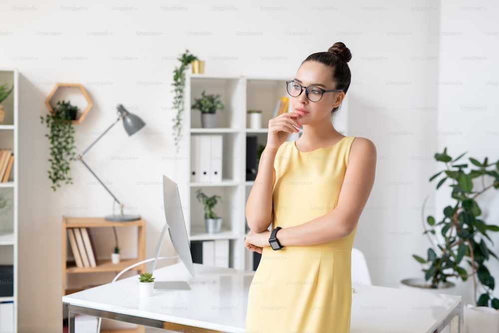 Pretty young pensive businesswoman in elegant yellow dress and eyeglasses standing by workplace in office