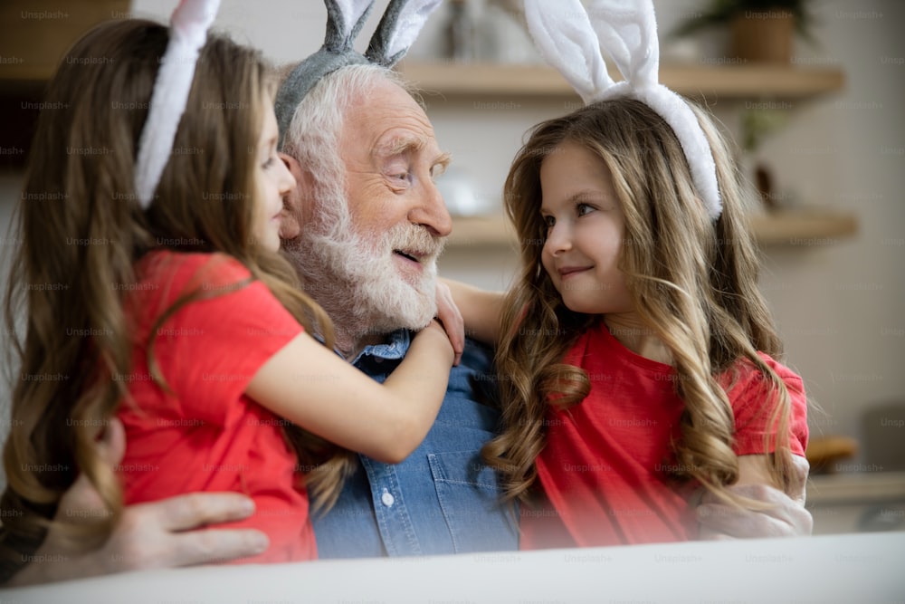 Mirthful aged man hugging his twin granddaughters and smiling to them
