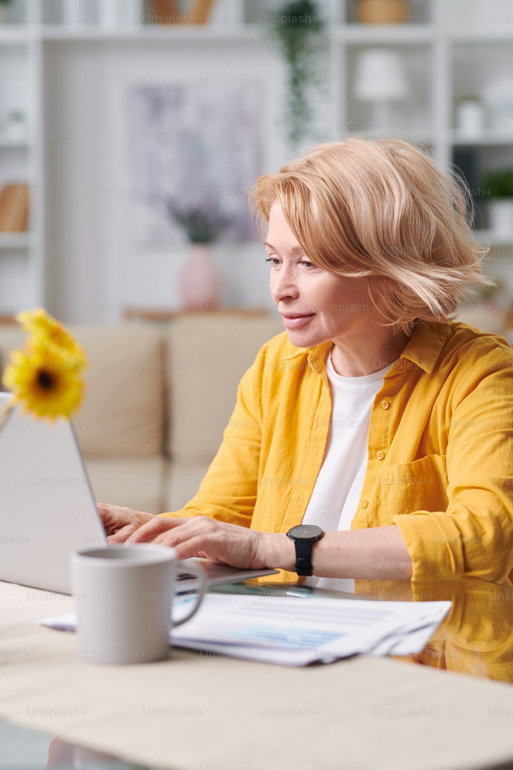 Pretty blond mature female in yellow jacket sitting by desk in front of laptop while networking in home environment
