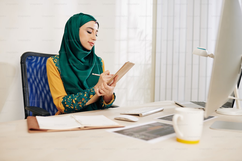 Imprenditrice musulmana seria seduta alla sua scrivania e prendendo appunti in agenda