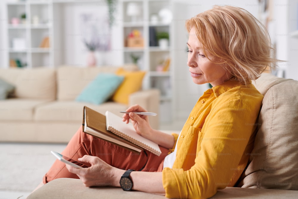 Pretty blond female in casualwear writing down plan of working day while sitting on sofa and scrolling in smartphone at home