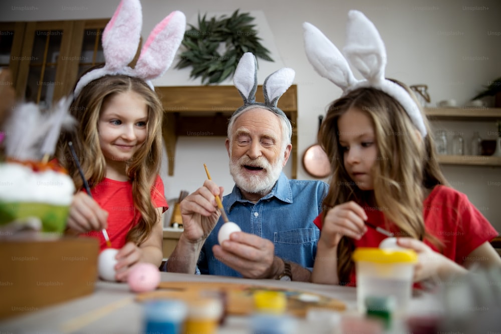 Hombre de edad positiva con orejas de conejo en la cabeza coloreando huevos de Pascua con nietas gemelas