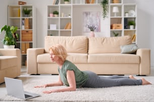 Mature attractive blond woman in activewear lying in front of laptop while repeating exercise after instructor during online fitness course